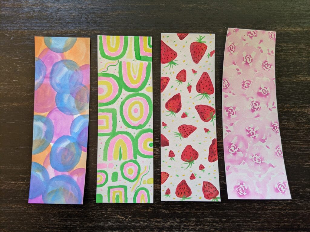 Four bookmarks with watercolor paintings. From left to right: translucent bubbles in blue, orange, and purple; abstract shapes in pastel pink, yellow, and green; ripe red strawberries with flecks of green and yellow in the background; soft purple pink flower petals. 